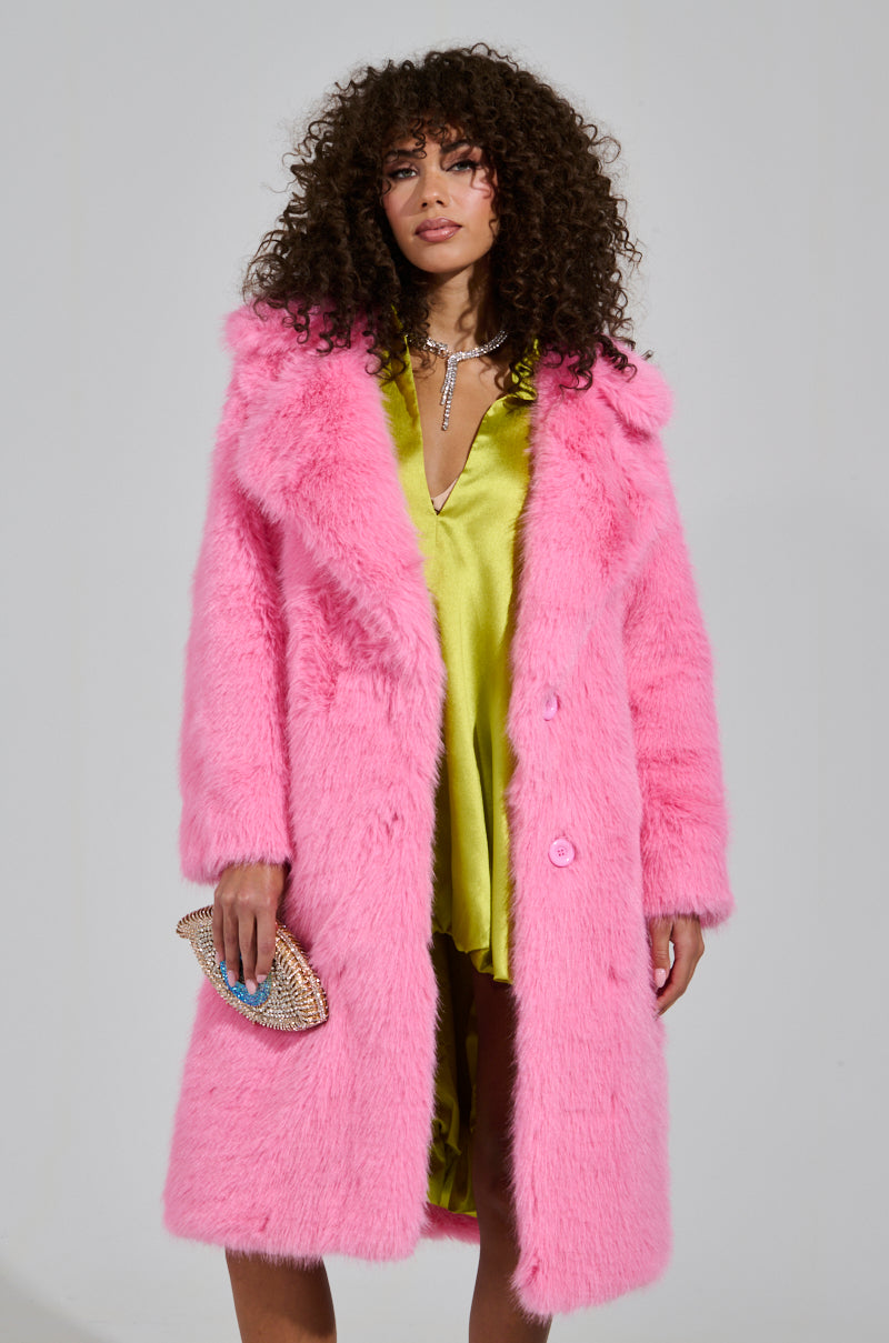 A person with curly hair captivates in the LENOX FAUX FUR TRENCH IN PINK layered over a yellow satin dress, holding a small sparkly clutch. The plain background emphasizes the high-quality faux fur and vibrant colors of the ensemble.