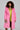 A person with curly hair captivates in the LENOX FAUX FUR TRENCH IN PINK layered over a yellow satin dress, holding a small sparkly clutch. The plain background emphasizes the high-quality faux fur and vibrant colors of the ensemble.