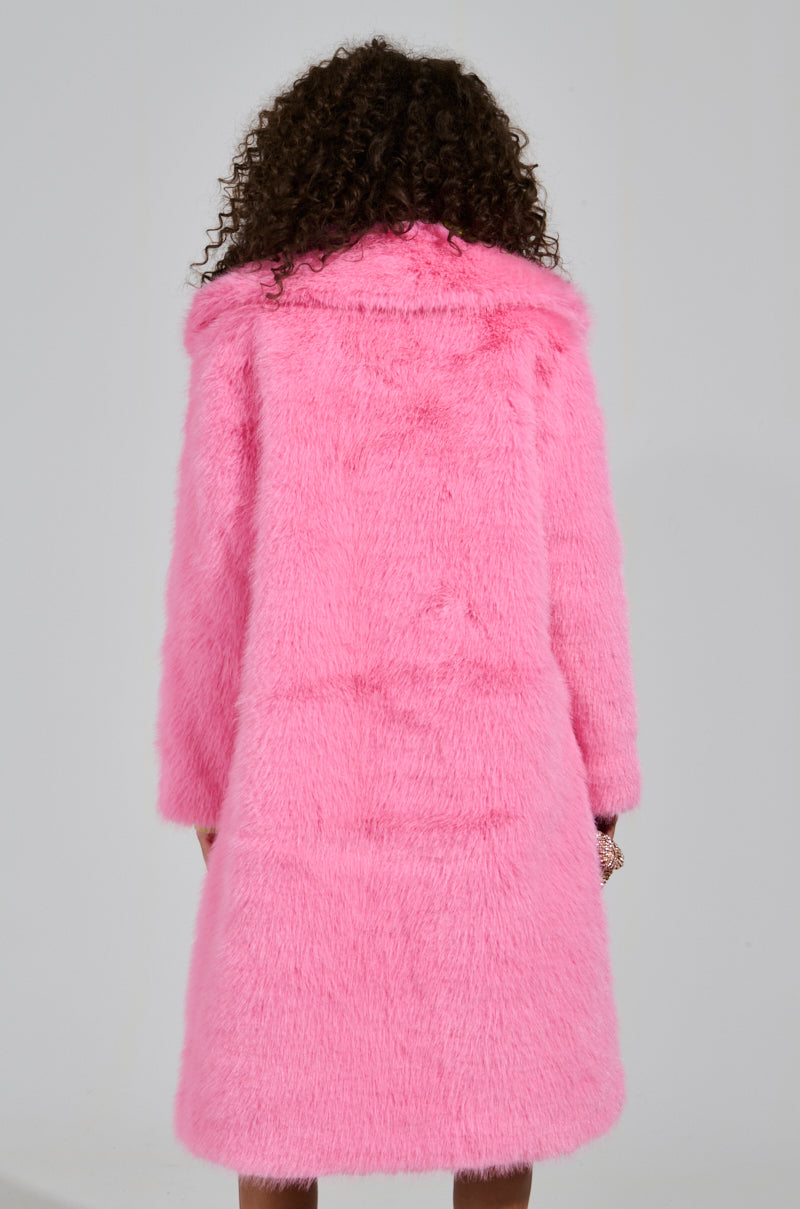 A person with curly hair is wearing the LENOX FAUX FUR TRENCH IN PINK, an oversized faux fur coat in a vibrant pink hue. They are facing away to highlight the back of this high-quality coat, which looks soft and plush against a plain, light-colored wall.