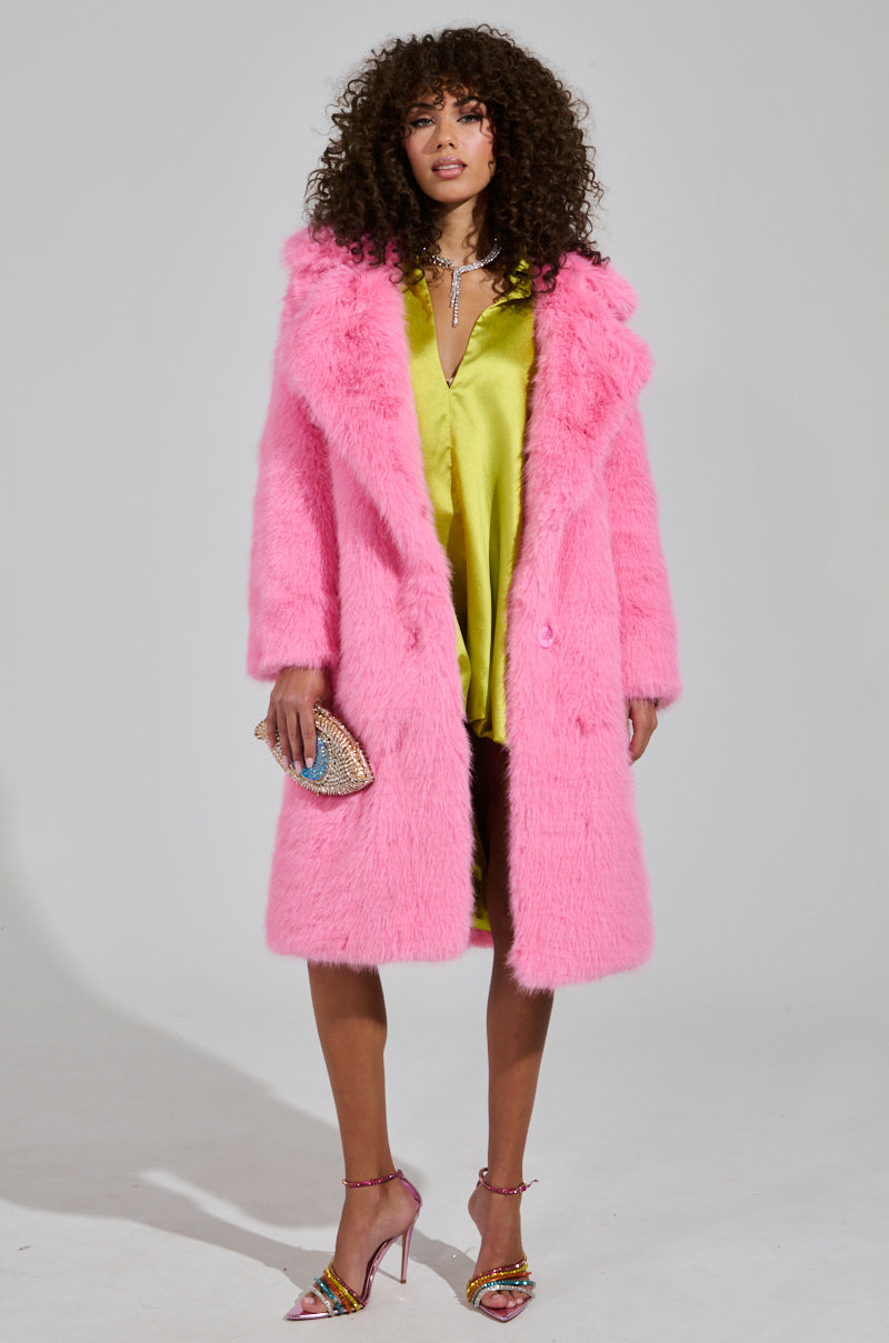 A woman confidently stands in the LENOX FAUX FUR TRENCH IN PINK, an oversized vibrant pink faux fur coat, layered over a bright yellow dress. She holds a sparkling clutch and dons colorful high heels. Her curly hair enhances the bold, fashionable look against a plain backdrop, perfectly highlighting the high-quality faux fur ensemble.