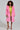 A woman confidently stands in the LENOX FAUX FUR TRENCH IN PINK, an oversized vibrant pink faux fur coat, layered over a bright yellow dress. She holds a sparkling clutch and dons colorful high heels. Her curly hair enhances the bold, fashionable look against a plain backdrop, perfectly highlighting the high-quality faux fur ensemble.