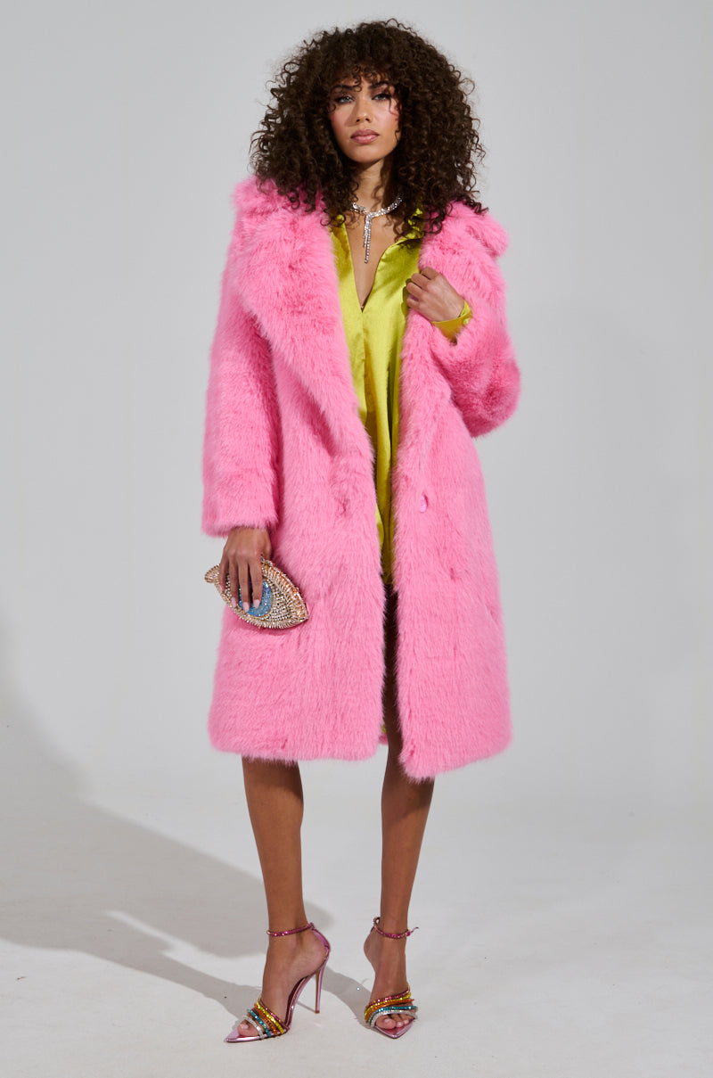 A woman stands confidently in the exquisite LENOX FAUX FUR TRENCH IN PINK over a striking lime green dress. She holds a sparkling clutch and wears high heels embellished with jewels. Her curly hair frames her face as she poses against a plain background, exuding elegance and style.