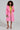 A woman stands confidently in the exquisite LENOX FAUX FUR TRENCH IN PINK over a striking lime green dress. She holds a sparkling clutch and wears high heels embellished with jewels. Her curly hair frames her face as she poses against a plain background, exuding elegance and style.