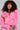 A person with curly hair is wearing the elegant LENOX FAUX FUR TRENCH IN PINK and holding a sparkling clutch. They have vibrant makeup and a jeweled necklace, standing against a plain background.