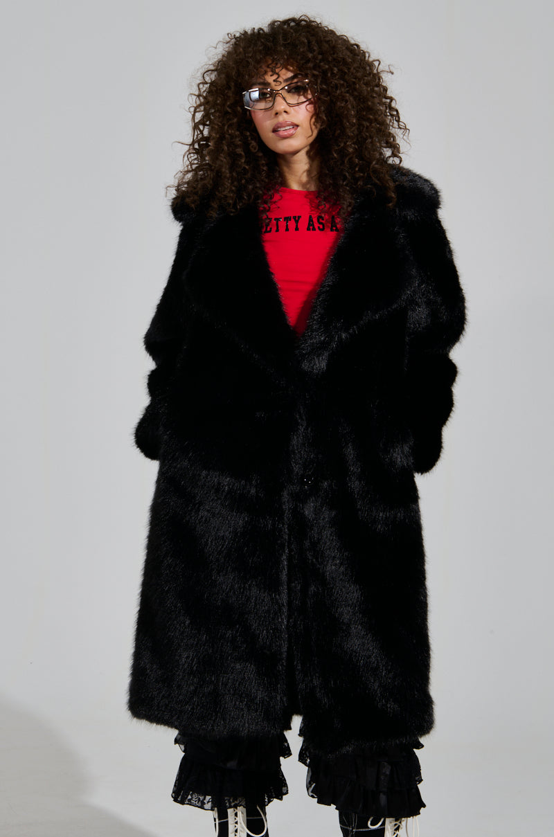 A person with curly hair wearing sunglasses, a red shirt, and a luxurious black LENOX FAUX FUR TRENCH. They also have black ruffled pants and white sneakers, standing against a plain background.