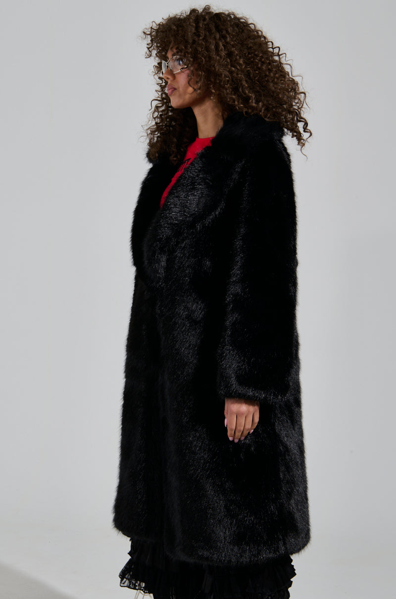 A person with curly hair stands in profile, draped in luxurious outerwear—a LENOX FAUX FUR TRENCH IN BLACK over a red top and black skirt. The background is plain and light-colored.