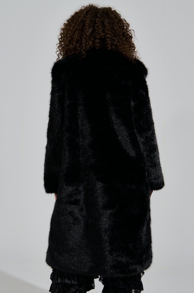 A person with curly hair is seen wearing the oversized LENOX FAUX FUR TRENCH IN BLACK, a luxurious outerwear garment, as they face away from the camera against a neutral background.