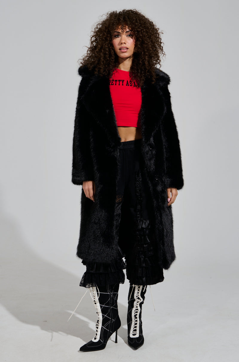 A woman stands confidently in the luxurious LENOX Faux Fur Trench in black, over a red cropped top with text. She pairs it with black pants featuring ruffled hems and white lace-up boots. With her curly hair cascading around her, she poses against a plain backdrop.