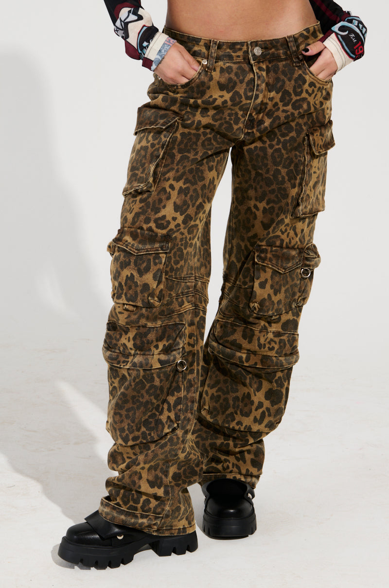 A person dressed in THE ULTIMATE LEOPARD CARGO PANT, featuring a mid-rise design, stands confidently with one hand on their hip, paired with black platform shoes. The relaxed silhouette against a plain white background exudes effortless style.