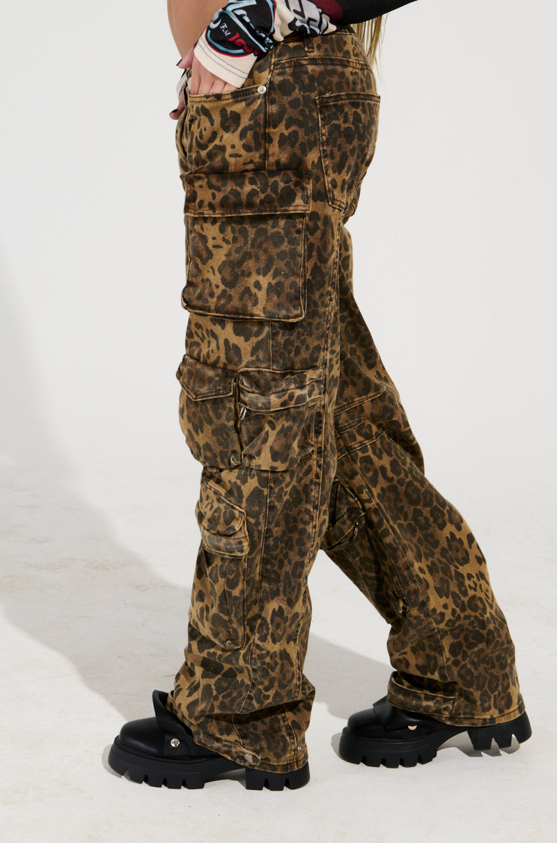 A person stands confidently against a plain background, wearing THE ULTIMATE LEOPARD CARGO PANT with a relaxed silhouette and multiple pockets, along with black chunky shoes. Their arms are crossed, perfectly showcasing the striking leopard print design of their outfit.