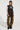 A person stands confidently wearing a sleeveless black top with text and fringe details, paired with THE ULTIMATE LEOPARD CARGO PANT in a relaxed silhouette and black boots. They have braided hair and are posing against a plain white background.