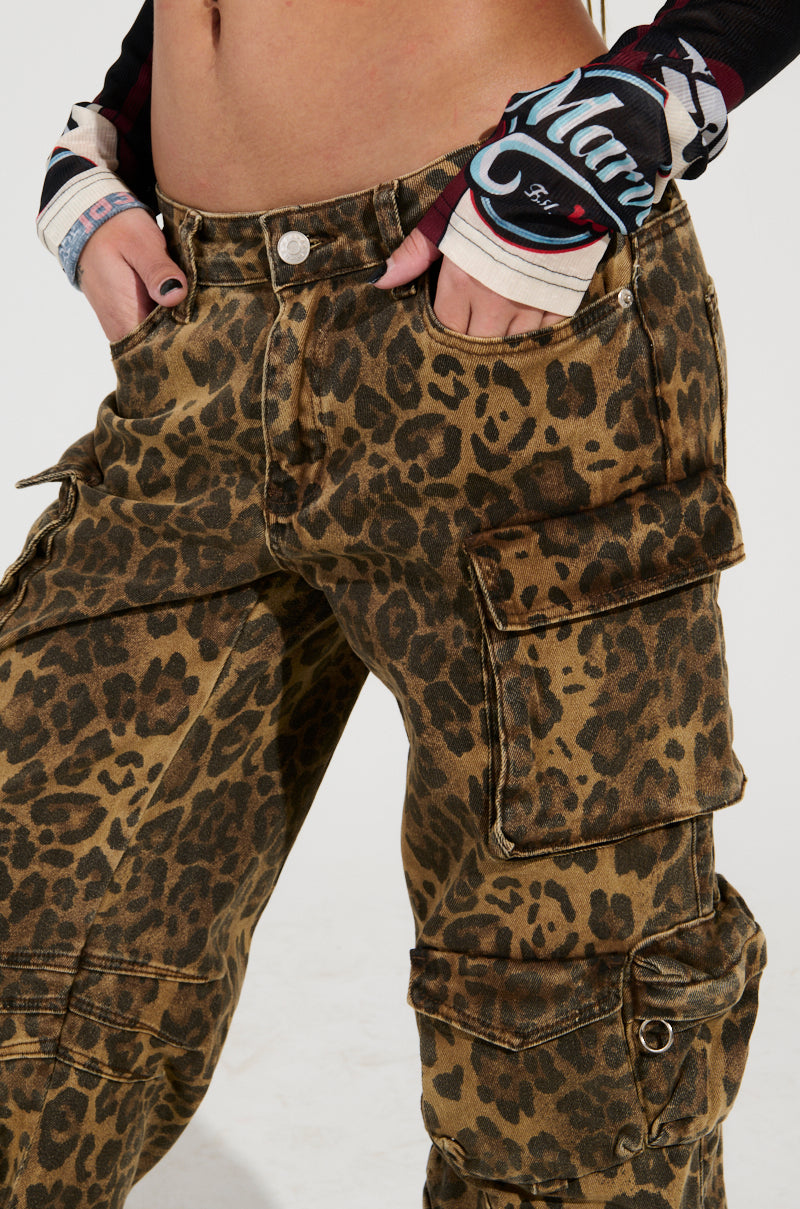 A person showcasing a relaxed silhouette in THE ULTIMATE LEOPARD CARGO PANT and a long-sleeve shirt with a tattoo-style design, hands casually tucked in pockets. The photo highlights the bold pattern of the pants and the laid-back pose.