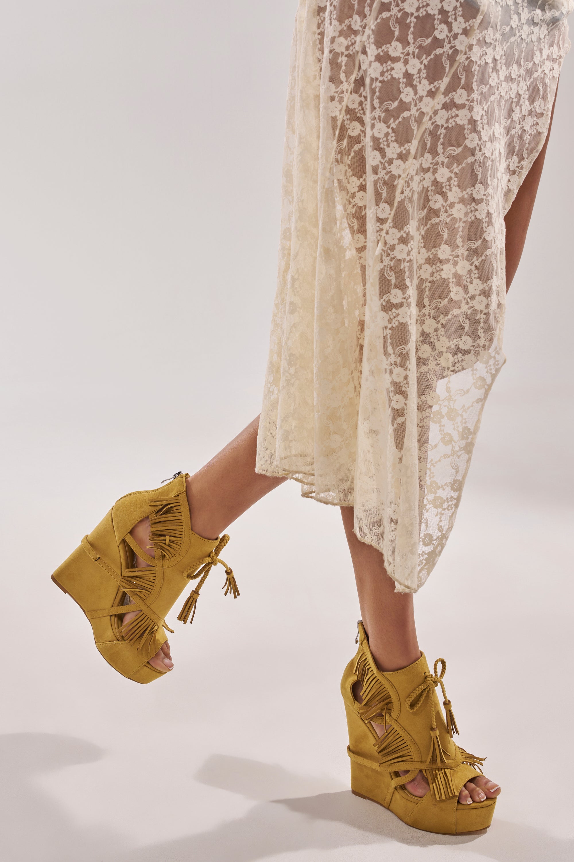 A person is mid-step in a sheer white lace skirt and AZALEA WANG LEONTINE Warm Olive Wedge Faux Suede Sandals against a plain background. 2