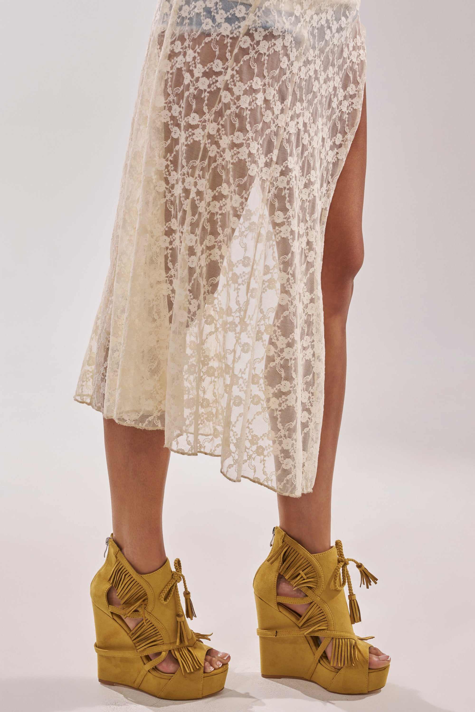 A person in a sheer cream lace skirt stands with one leg forward, showcasing the AZALEA WANG LEONTINE WARM OLIVE WEDGE FAUX SUEDE SANDAL. The sandals feature a platform sole and playful yellow tassel fringe. The background is light and plain. 7