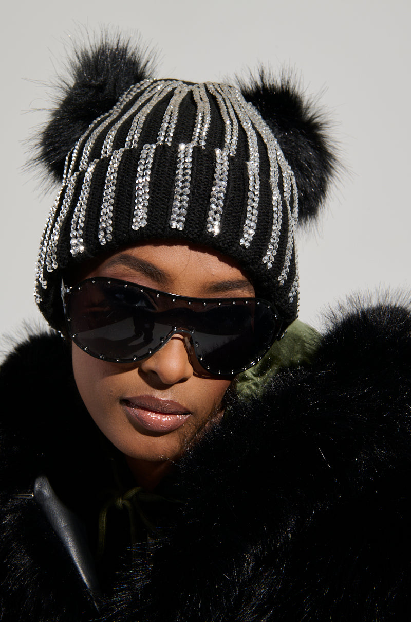A person wears large sunglasses and a cozy black fur coat, perfectly paired with the chic LEXII Rhinestone Knit Mouse Beanie in Black, adorned with rhinestones and playful pom-poms on top. The background remains plain and light-colored.