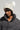 A person wearing a black, textured jacket and the LEXII Rhinestone Knit Mouse Beanie in White, adorned with mouse ear accents, gazes directly at the camera. The beanie showcases black stripes and a fluffy faux fur pom-pom, accentuating their neutral expression against a plain background.