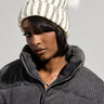 A person wearing a black, textured jacket and the LEXII Rhinestone Knit Mouse Beanie in White, adorned with mouse ear accents, gazes directly at the camera. The beanie showcases black stripes and a fluffy faux fur pom-pom, accentuating their neutral expression against a plain background.
