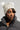 A person wearing the LEXII Rhinestone Knit Mouse Beanie in White, adorned with chic pom-poms, pairs it with a quilted dark jacket. Their head is slightly turned to the side, highlighting their makeup and the intricate texture of the jacket against a neutral gray background.