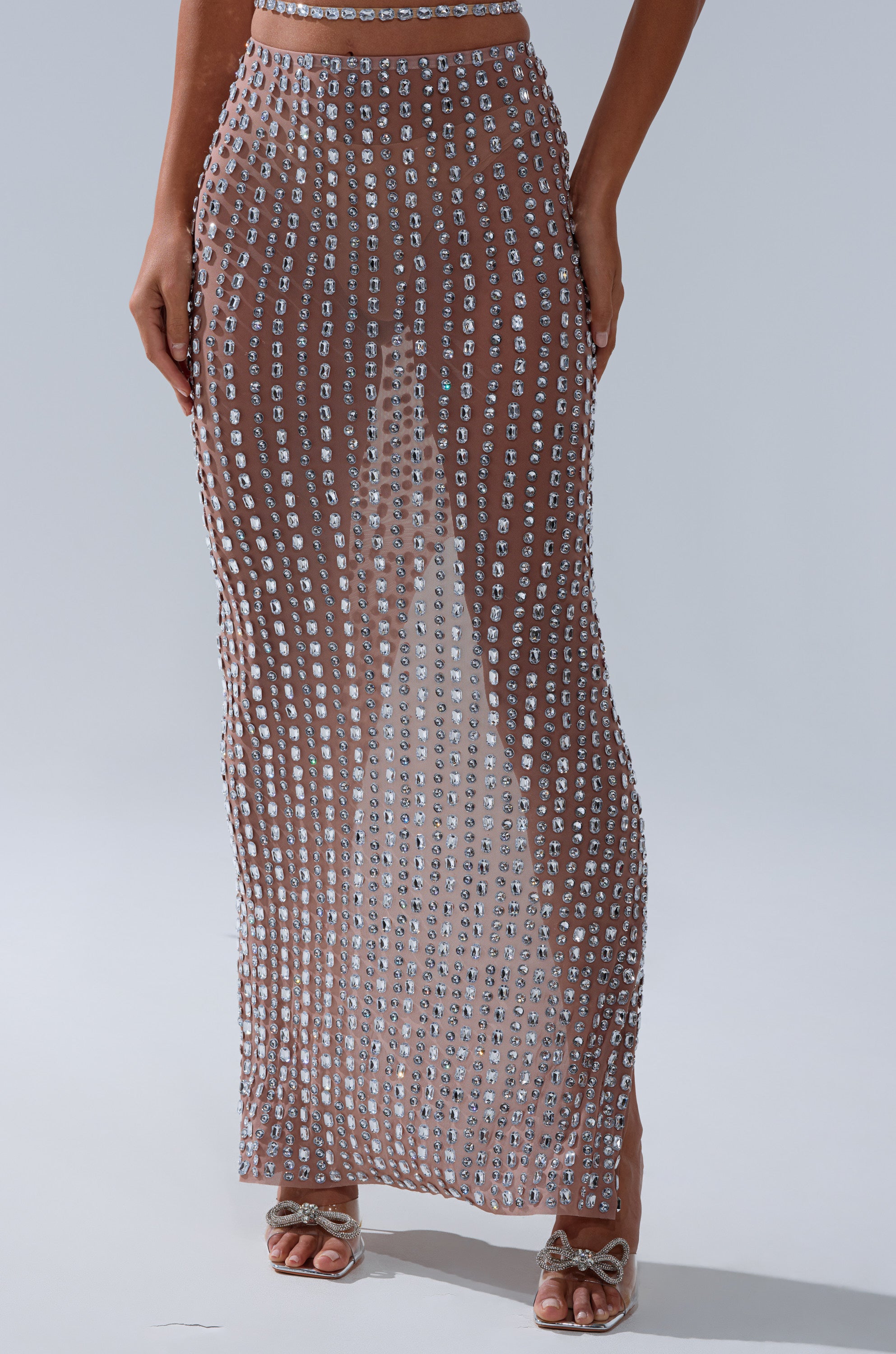 A person wears the ALL THE SHINE RHINESTONE MAXI SKIRT, a long sheer maxi skirt embellished with sparkling squares, paired with strappy heeled sandals against a plain, light background.