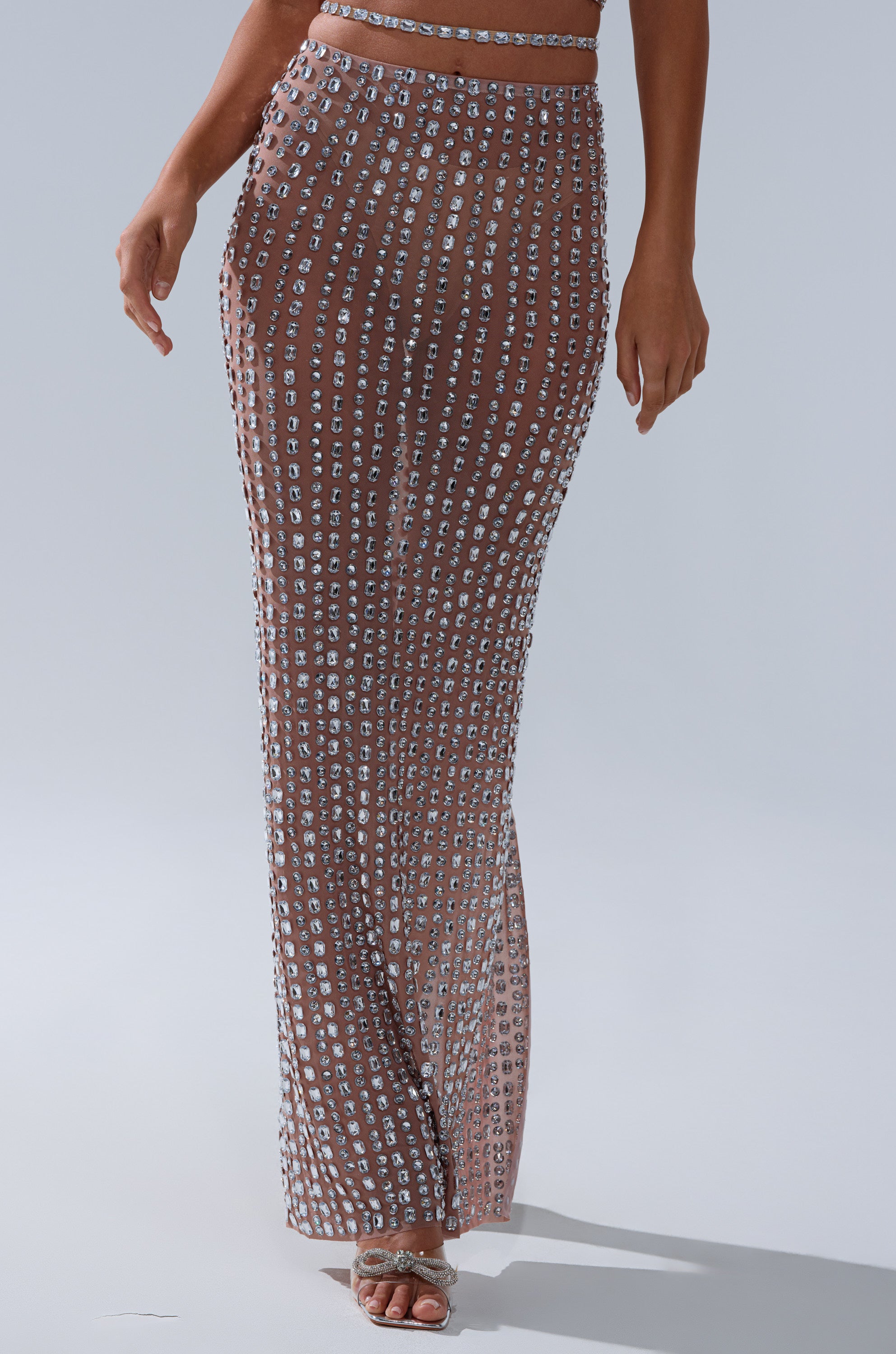 A person models the ALL THE SHINE RHINESTONE MAXI SKIRT, featuring vertical rows of silver rhinestones, styled with open-toe heels against a plain light background.