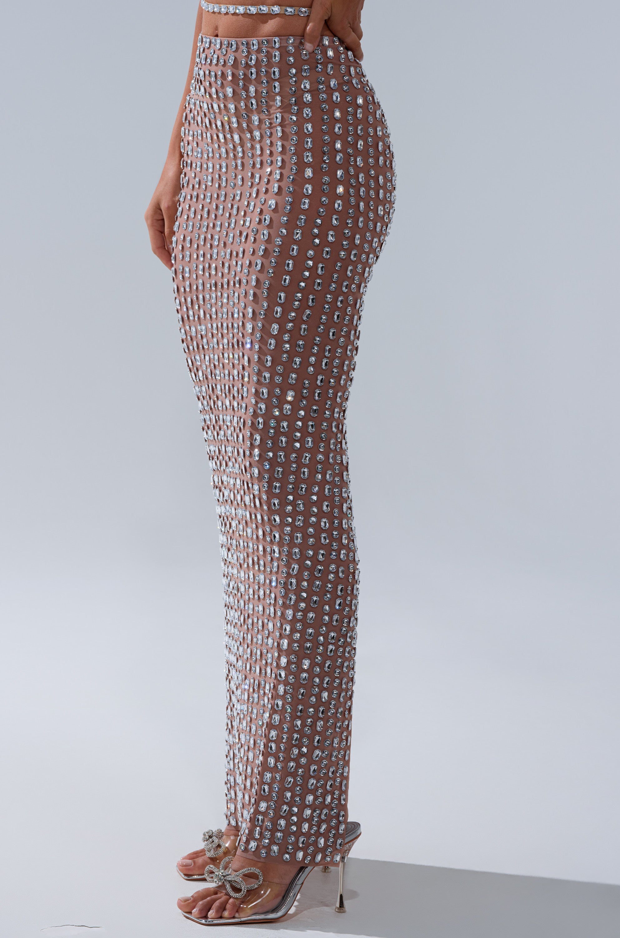 A person stands wearing the ALL THE SHINE RHINESTONE MAXI SKIRT, a sheer embellished maxi skirt with sparkling square accents, paired with rhinestone-embellished high-heeled sandals against a plain light gray background.