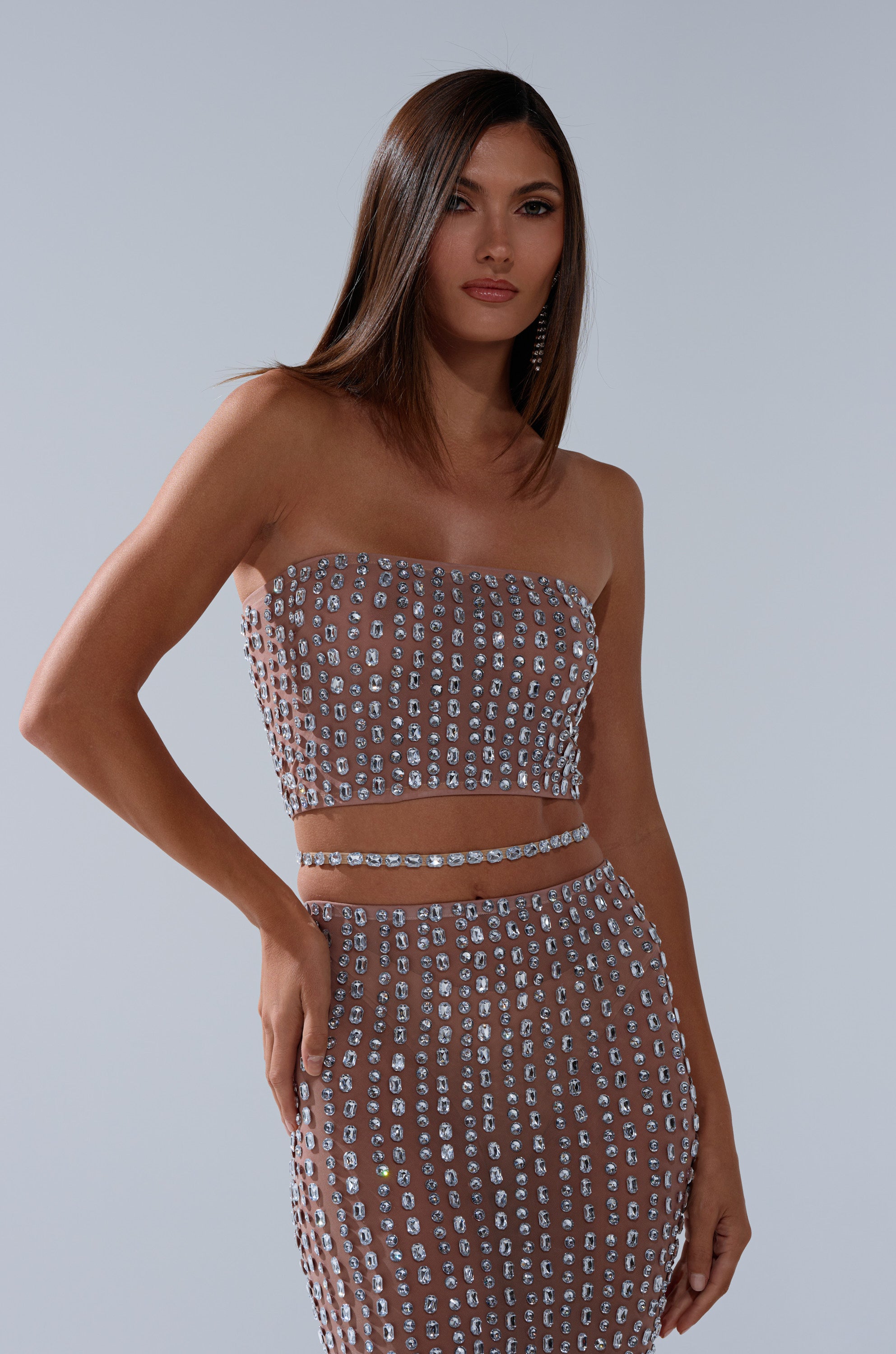 A woman with straight brown hair wears the ALL THE SHINE RHINESTONE BANDEAU and matching skirt, both featuring a grid of sparkling square embellishments, against a plain light background.