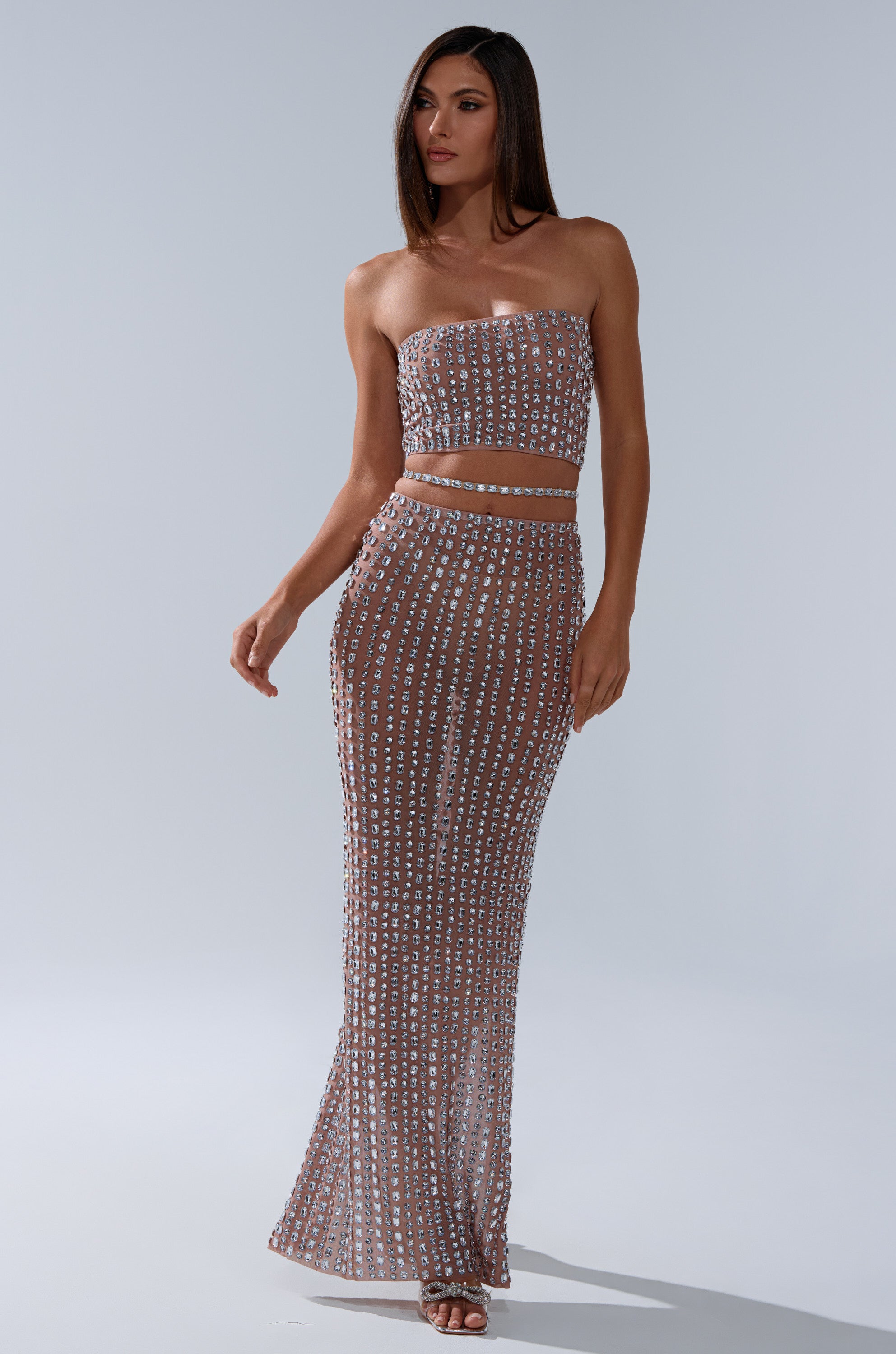 A woman wearing the ALL THE SHINE RHINESTONE BANDEAU and a matching high-waisted, floor-length skirt with sparkling accents poses against a plain light gray background.