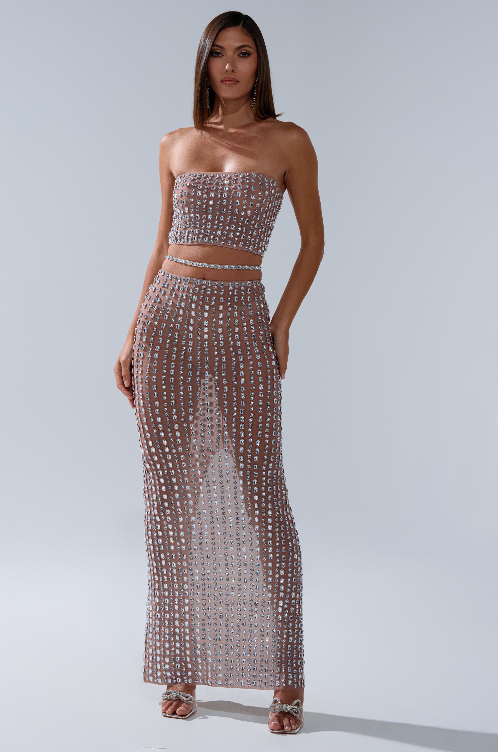 A woman models the ALL THE SHINE RHINESTONE BANDEAU, a sheer two-piece set with a sparkling bandeau top and long high-waisted skirt. She pairs the look with straight hair and open-toed heels.