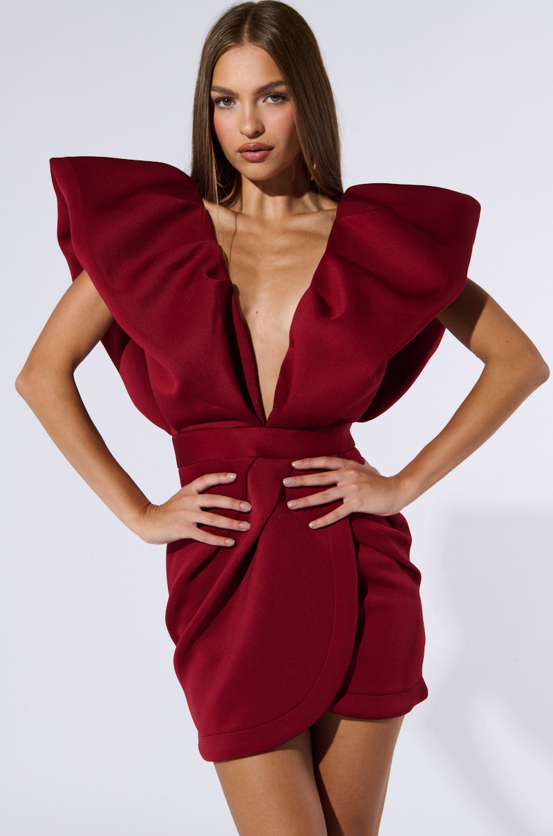 A woman stands confidently, wearing an IRREPLACEABLE RUFFLE SLEEVE TULIP HEM MINI DRESS IN BURGUNDY with large ruffle sleeves and a plunging neckline. Her hands rest on her hips as she gazes forward. The dress is form-fitting and short, highlighting her slender figure.