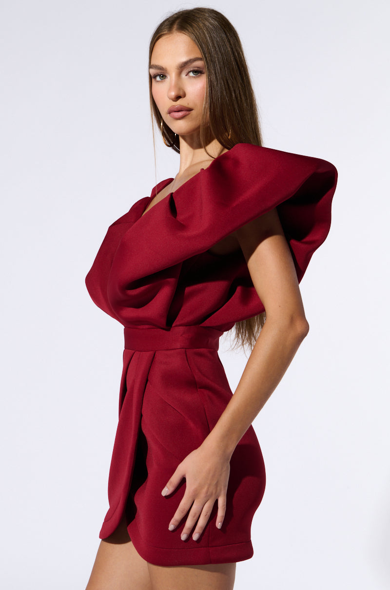 A woman with long brown hair is wearing the IRREPLACEABLE RUFFLE SLEEVE TULIP HEM MINI DRESS IN BURGUNDY, which features stylish ruffled details on one shoulder and a cinched waist. She poses confidently against a plain white background.