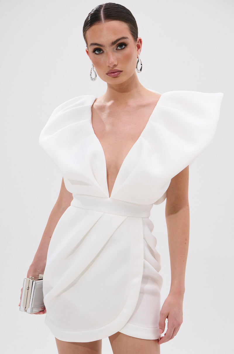 Wearing the IRREPLACEABLE RUFFLE SLEEVE TULIP HEM MINI DRESS IN WHITE featuring sculpted shoulders, a deep V neckline, and tulip hem, she carries a silver clutch, has slicked-back hair, and wears large hoop earrings.
