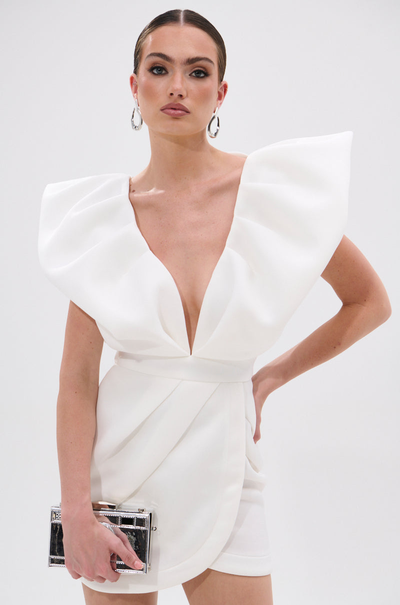 A woman poses against a plain background wearing the IRREPLACEABLE RUFFLE SLEEVE TULIP HEM MINI DRESS IN WHITE, featuring dramatic ruffle sleeves, a deep V-neckline, and a tulip hem. She carries a metallic clutch and wears hoop earrings with slicked-back hair.