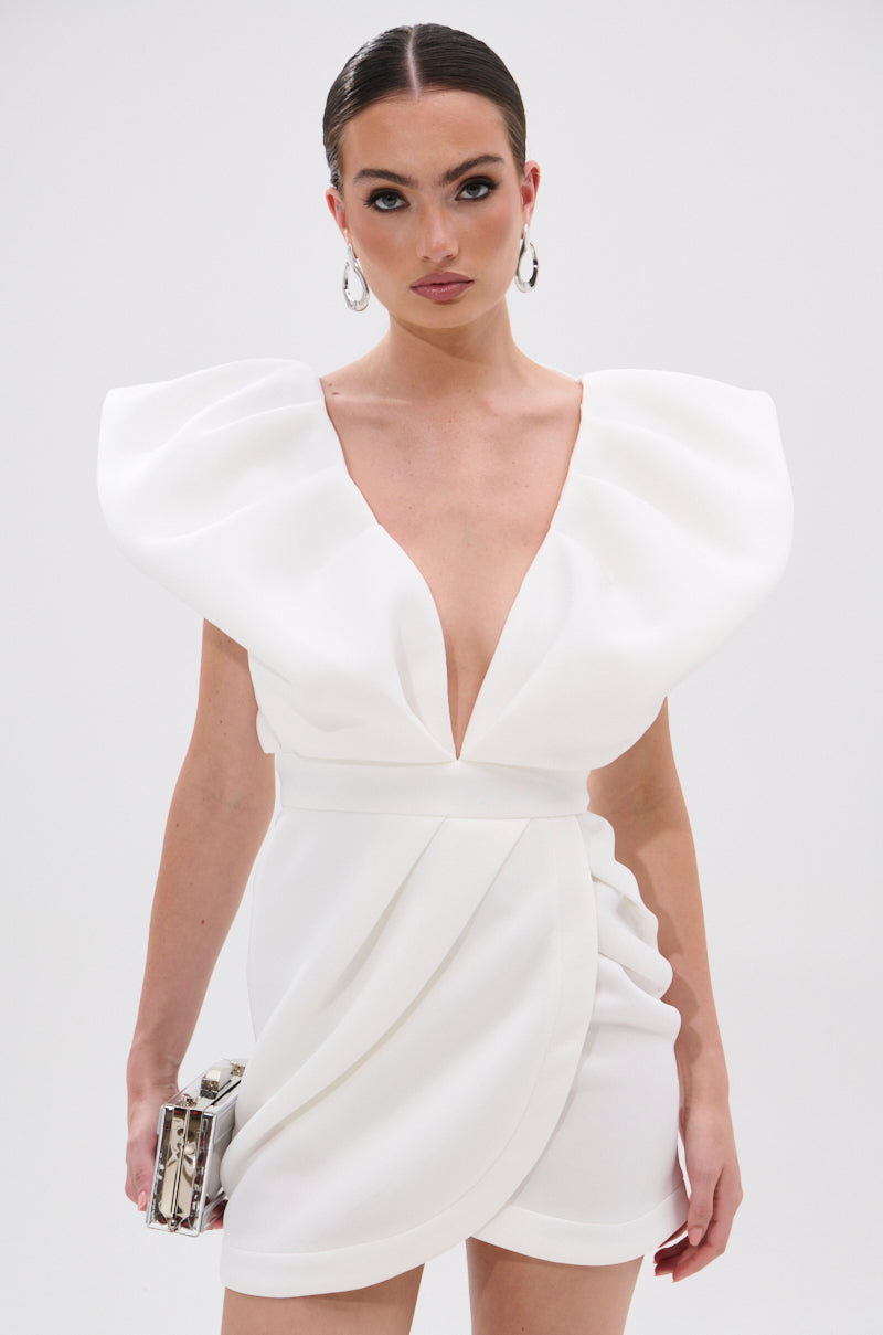 A woman in the IRREPLACEABLE RUFFLE SLEEVE TULIP HEM MINI DRESS IN WHITE with dramatic shoulders poses against a plain background, holding a metallic clutch and wearing hoop earrings.