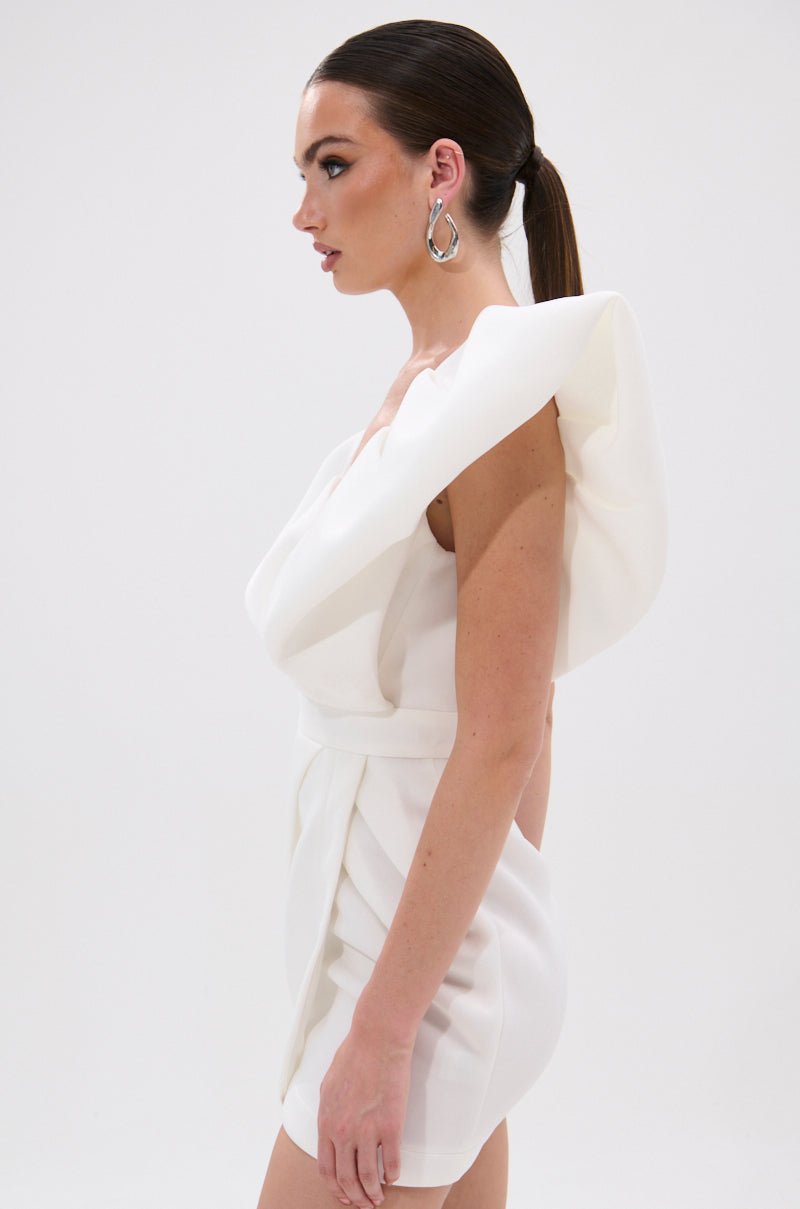 A woman stands in profile against a white background, wearing the IRREPLACEABLE RUFFLE SLEEVE TULIP HEM MINI DRESS IN WHITE with dramatic ruffle sleeves. Her hair is slicked back in a ponytail, and she accessorizes with large silver hoop earrings.