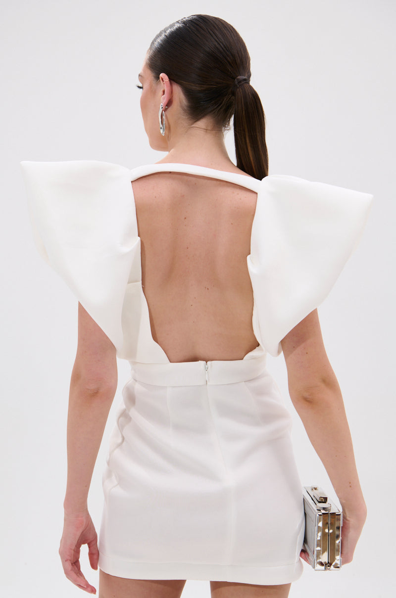 A woman with dark hair in a low ponytail wears the IRREPLACEABLE RUFFLE SLEEVE TULIP HEM MINI DRESS IN WHITE. Photographed from behind against a plain background, she holds a metallic clutch.