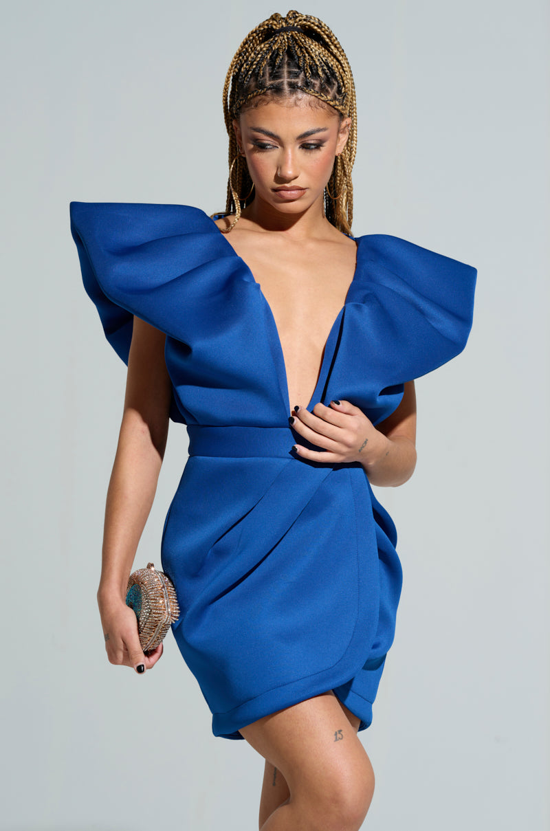 A person with braided hair wears the IRREPLACEABLE RUFFLE SLEEVE TULIP HEM MINI DRESS IN BLUE, featuring dramatic shoulders and a deep V-neckline. They hold a small textured clutch, the dress's tulip hem adding flair as they stand against a plain background.