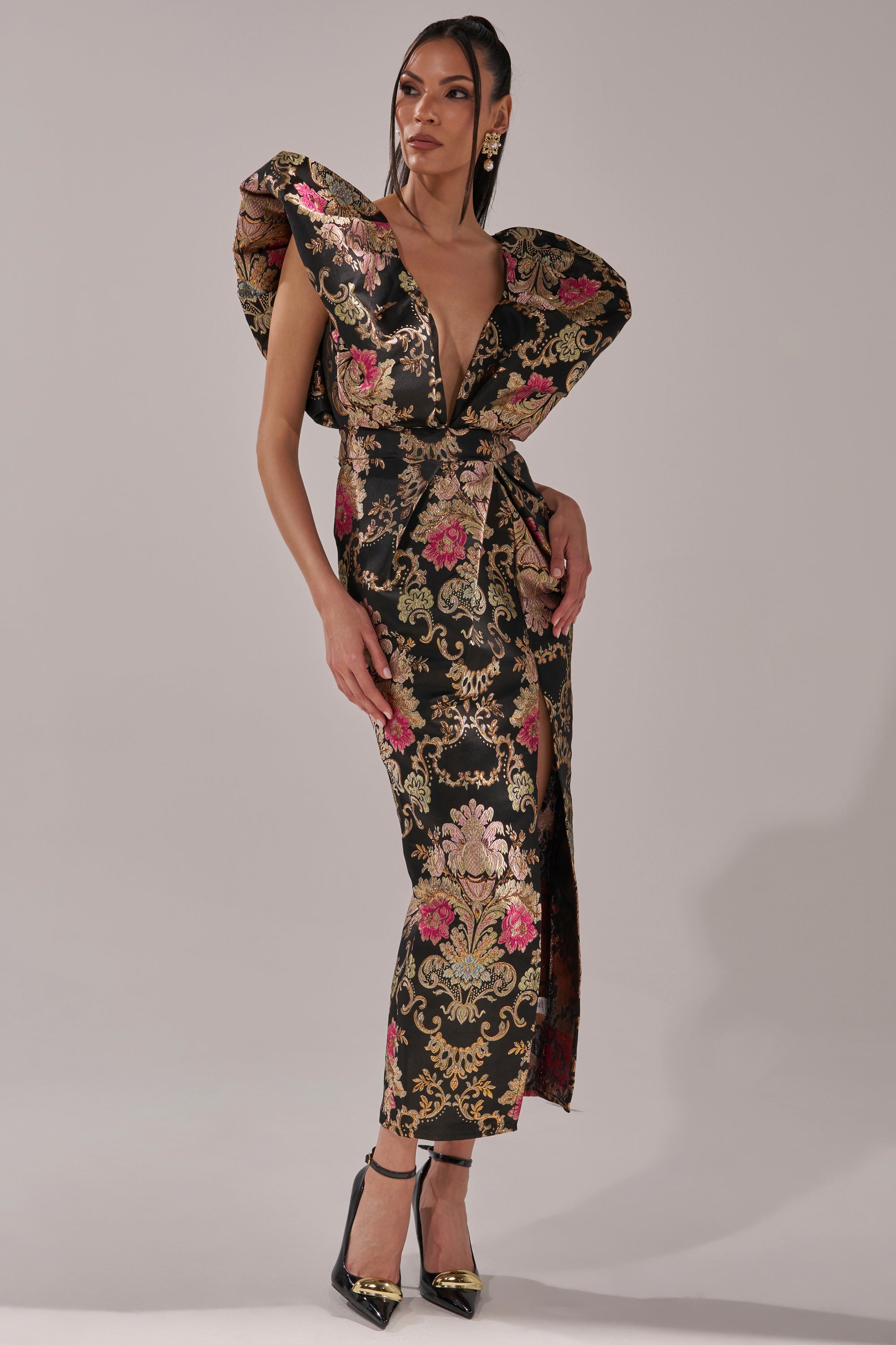 A woman poses in the BEST DRESSED BROCADE MAXI DRESS, featuring metallic gold, pink, and black floral brocade, oversized structured shoulders, a plunging neckline, and a high slit. She completes her bold look with high heels against a plain background.