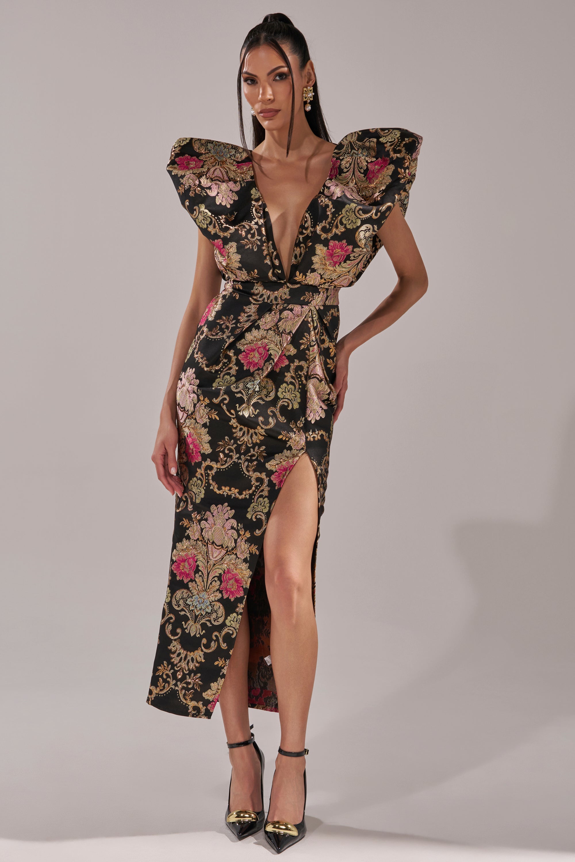 A woman models the BEST DRESSED BROCADE MAXI DRESS, a black metallic brocade gown with gold and pink florals, deep V-neck, bold shoulders, and a high slit. She completes the look with black heels and matching earrings against a plain backdrop.