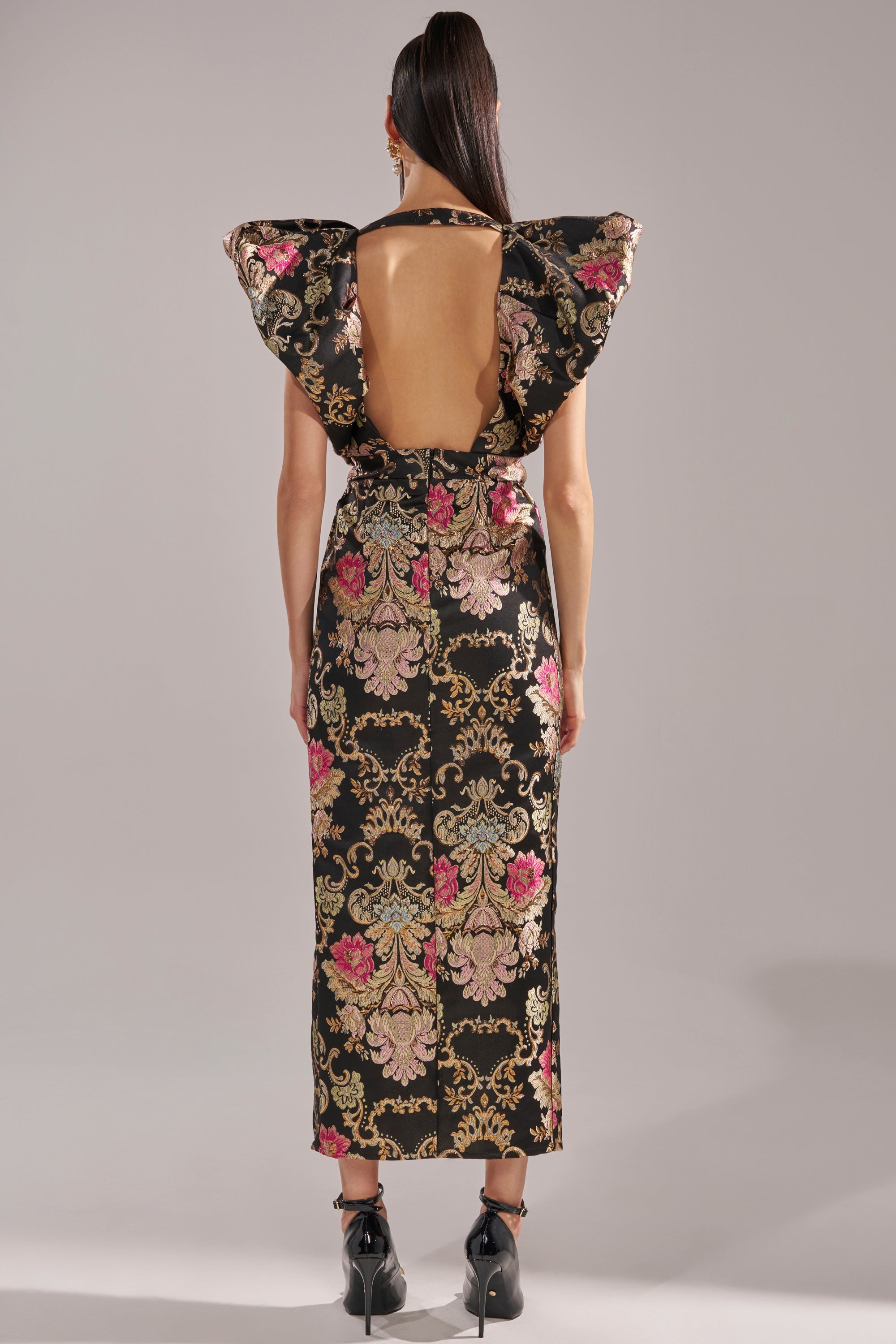 A person with long hair in a ponytail wears the BEST DRESSED BROCADE MAXI DRESS, featuring a black metallic brocade fabric, open back, bold ruffled shoulders, and pink and gold ornate floral pattern, paired with black high heels.