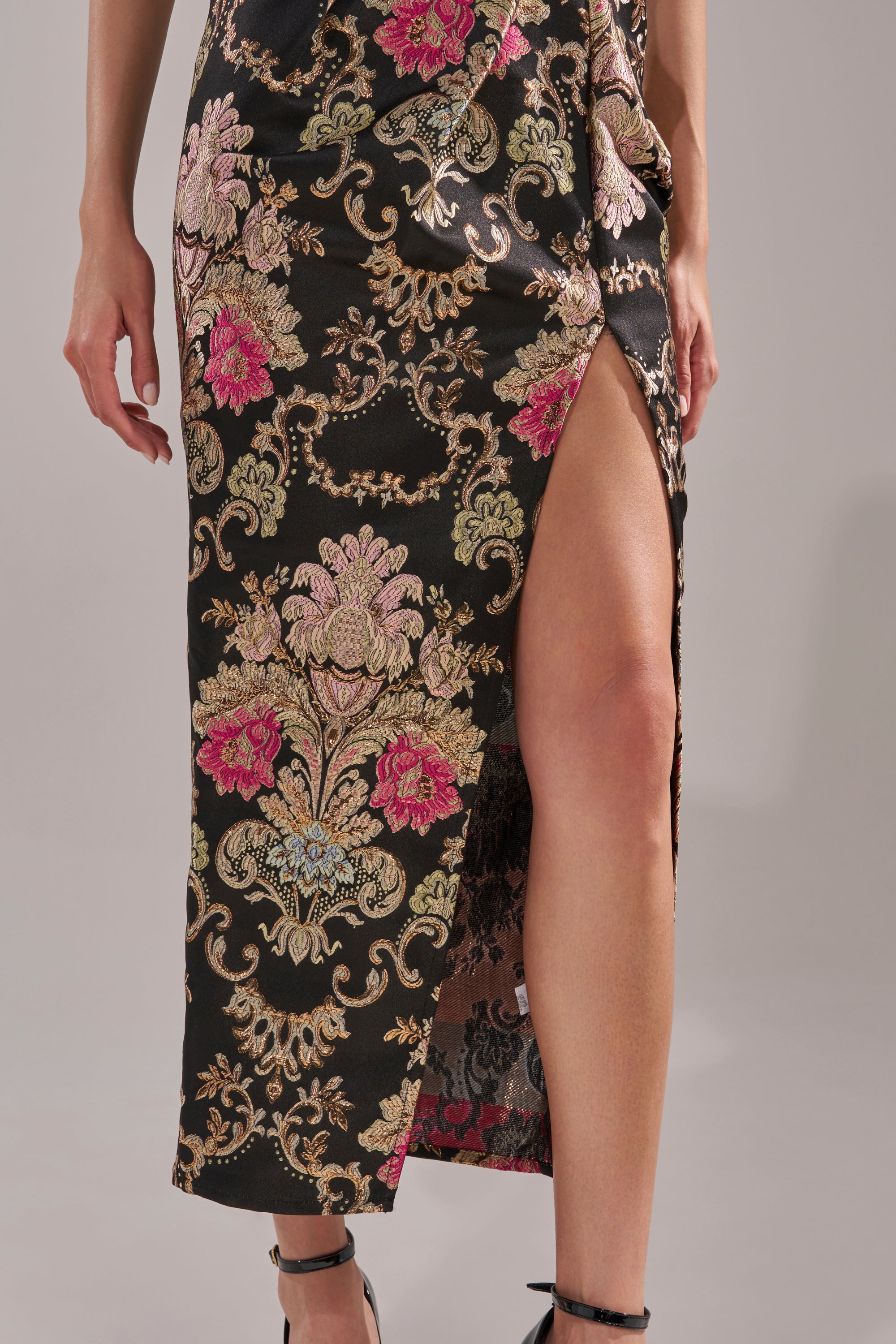 Wearing the BEST DRESSED BROCADE MAXI DRESS, featuring black metallic fabric with ornate pink, gold, and green floral patterns and a high side slit, paired with elegant black high-heeled shoes.