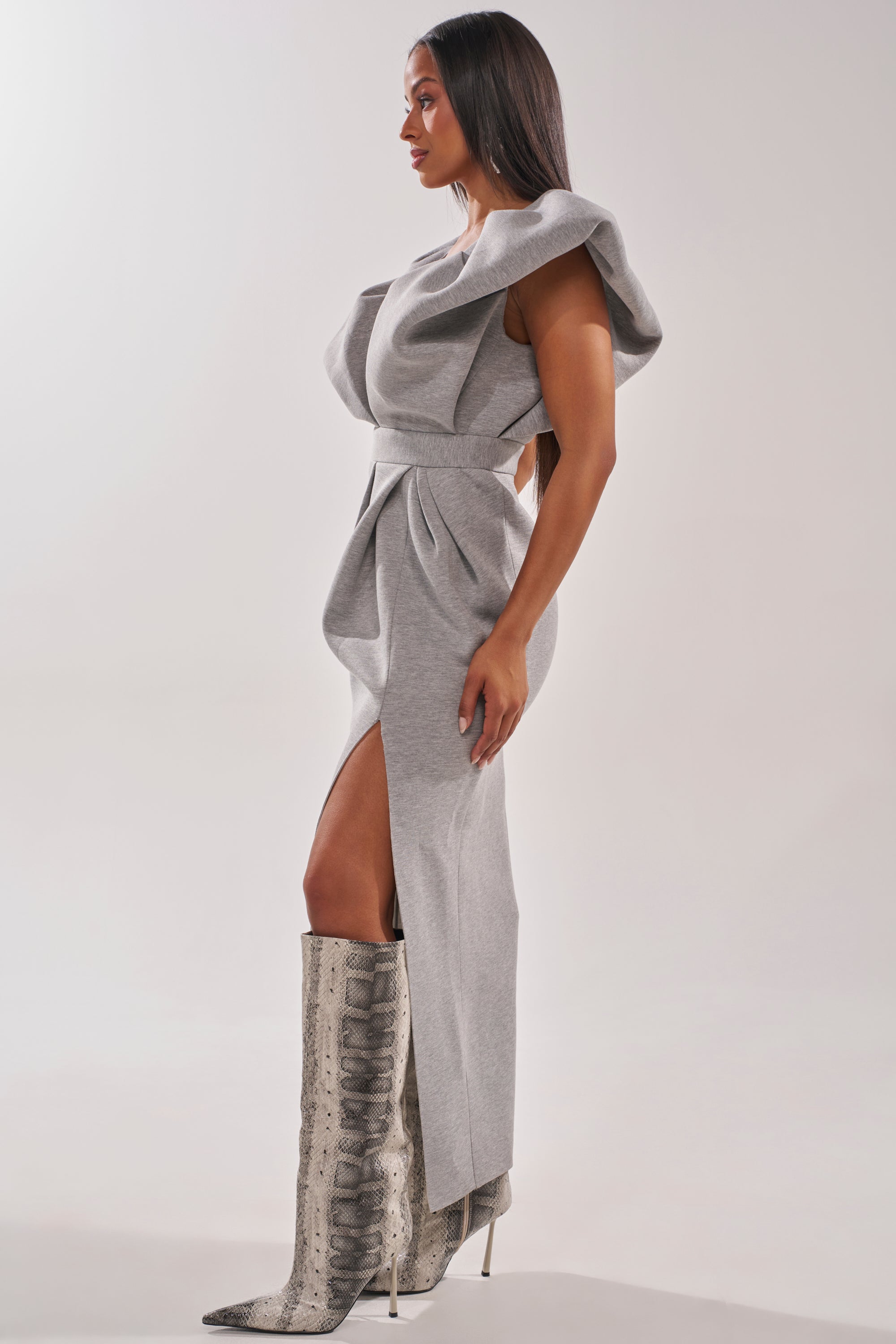 A woman stands sideways wearing the MAKE YOU MINE MAXI DRESS, a light gray, sculpted asymmetrical maxi with dramatic ruffles, architectural shoulders, and a high slit, styled with tall pointed snakeskin boots against a plain background. 1