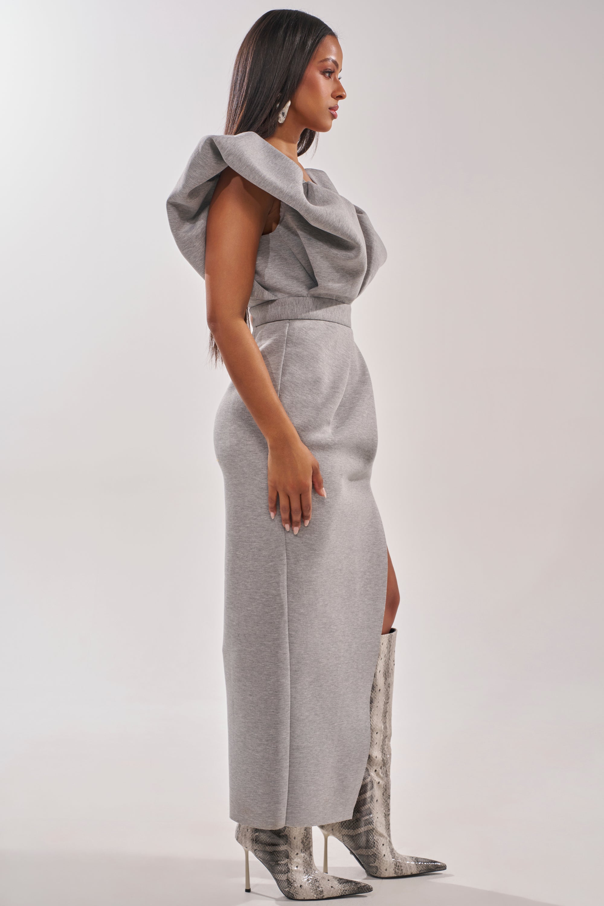 A woman stands in profile wearing the MAKE YOU MINE MAXI DRESS, a fitted, sleeveless gray dress with architectural shoulders and dramatic ruffled details, paired with knee-high snakeskin boots. The background is plain and light-colored. 2