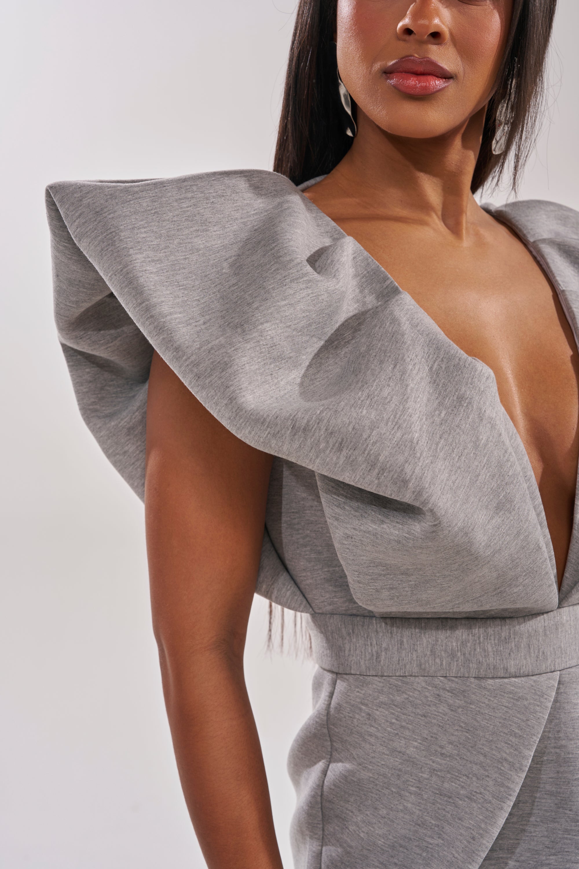 A woman wears the MAKE YOU MINE MAXI DRESS, a grey structured maxi with a deep V-neck, bold ruffled sleeves, and architectural shoulders, posing against a plain light background. 4