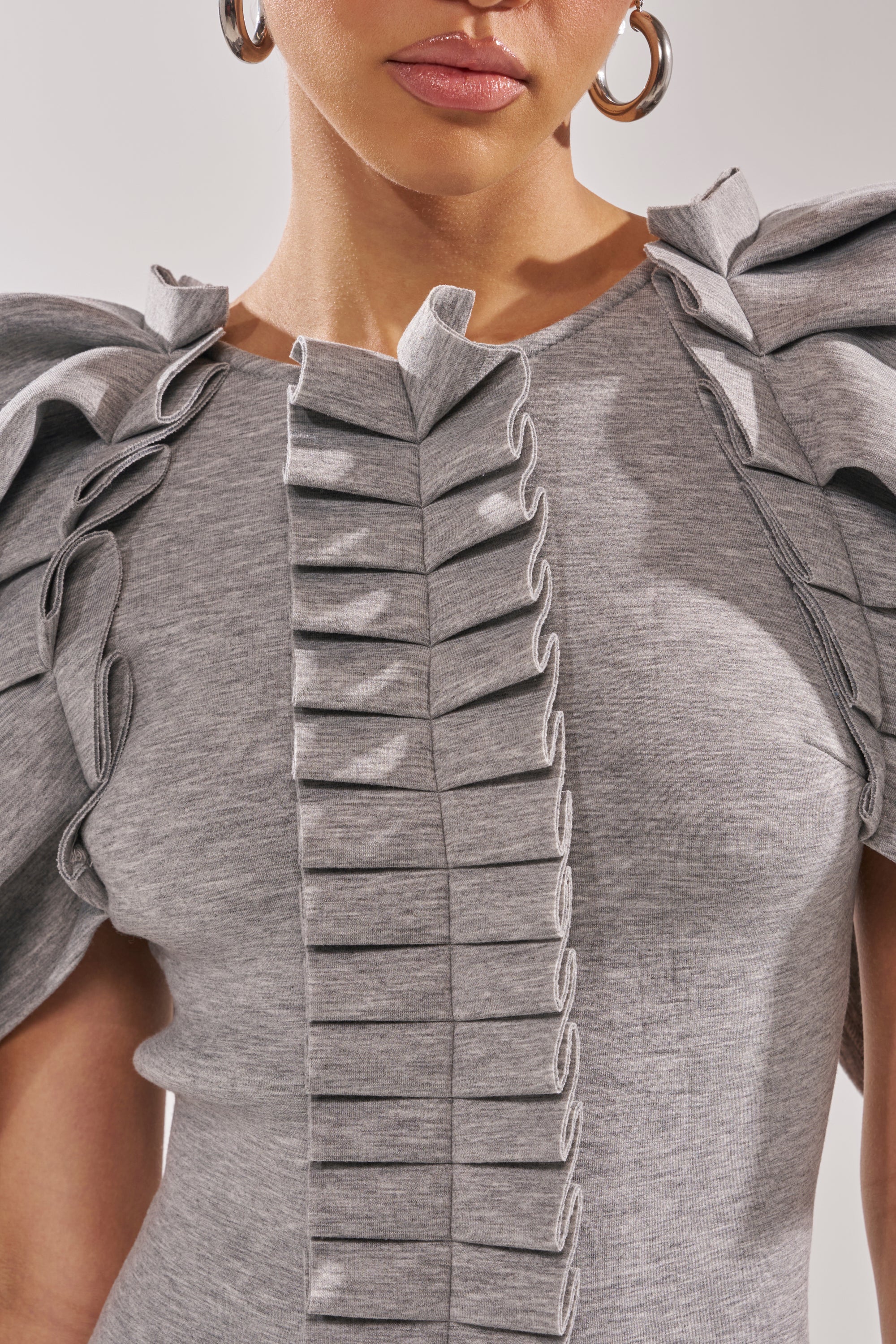 A woman models the SPREAD MY WINGS MINI DRESS in heather grey, featuring structured layered ruffle sleeves and a pleated front design, paired with hoop earrings. The image frames her from the shoulders to just above the mouth.