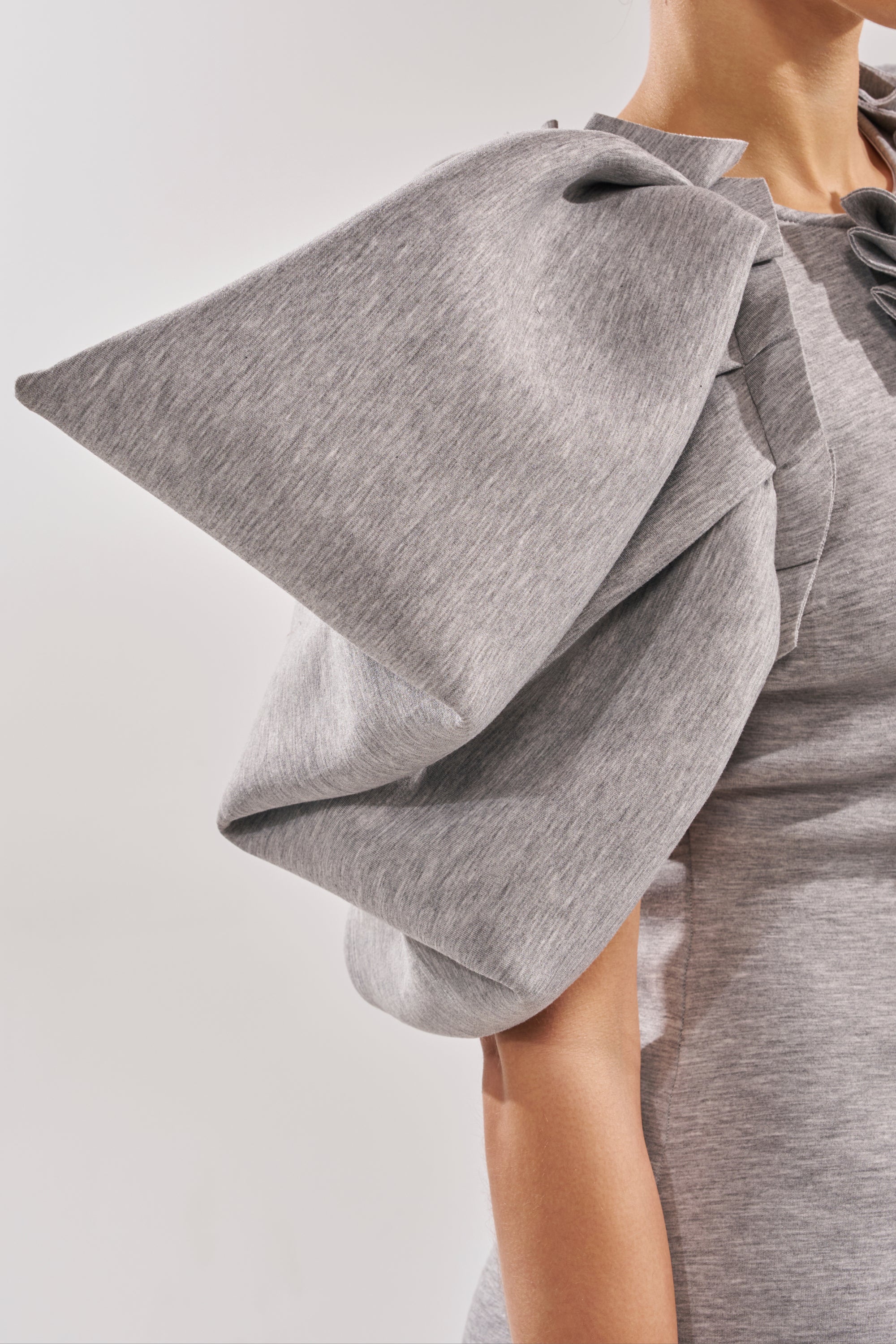 A close-up of a person wearing the SPREAD MY WINGS MINI DRESS in heather grey, featuring a bold, sculptural ruffle sleeve with dramatic, origami-like shaping. The background is plain and neutral.