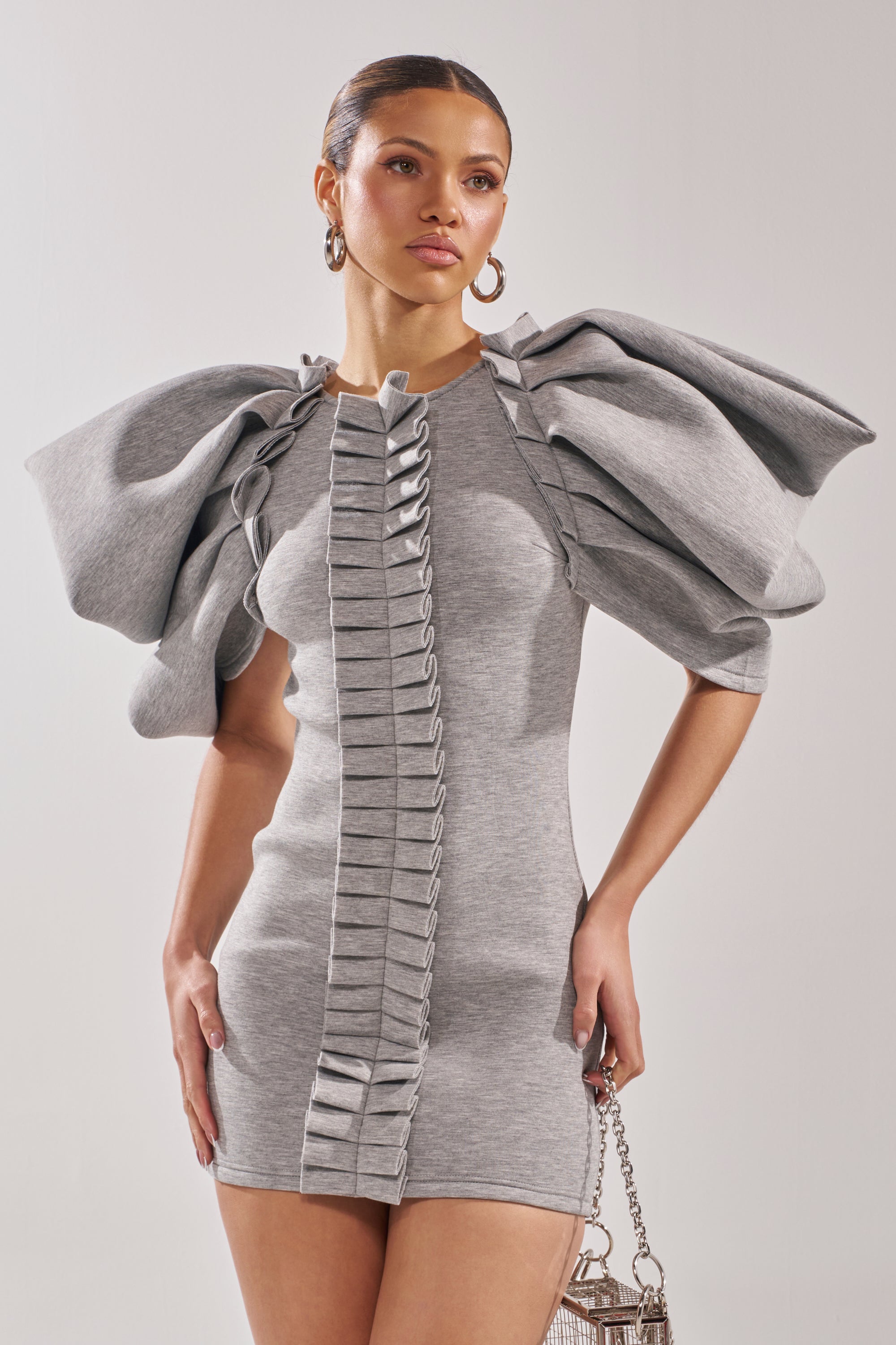 A woman wears the SPREAD MY WINGS MINI DRESS, a short heather grey dress with dramatic front ruffles. She holds a small silver handbag and stands against a plain background, her hair slicked back.
