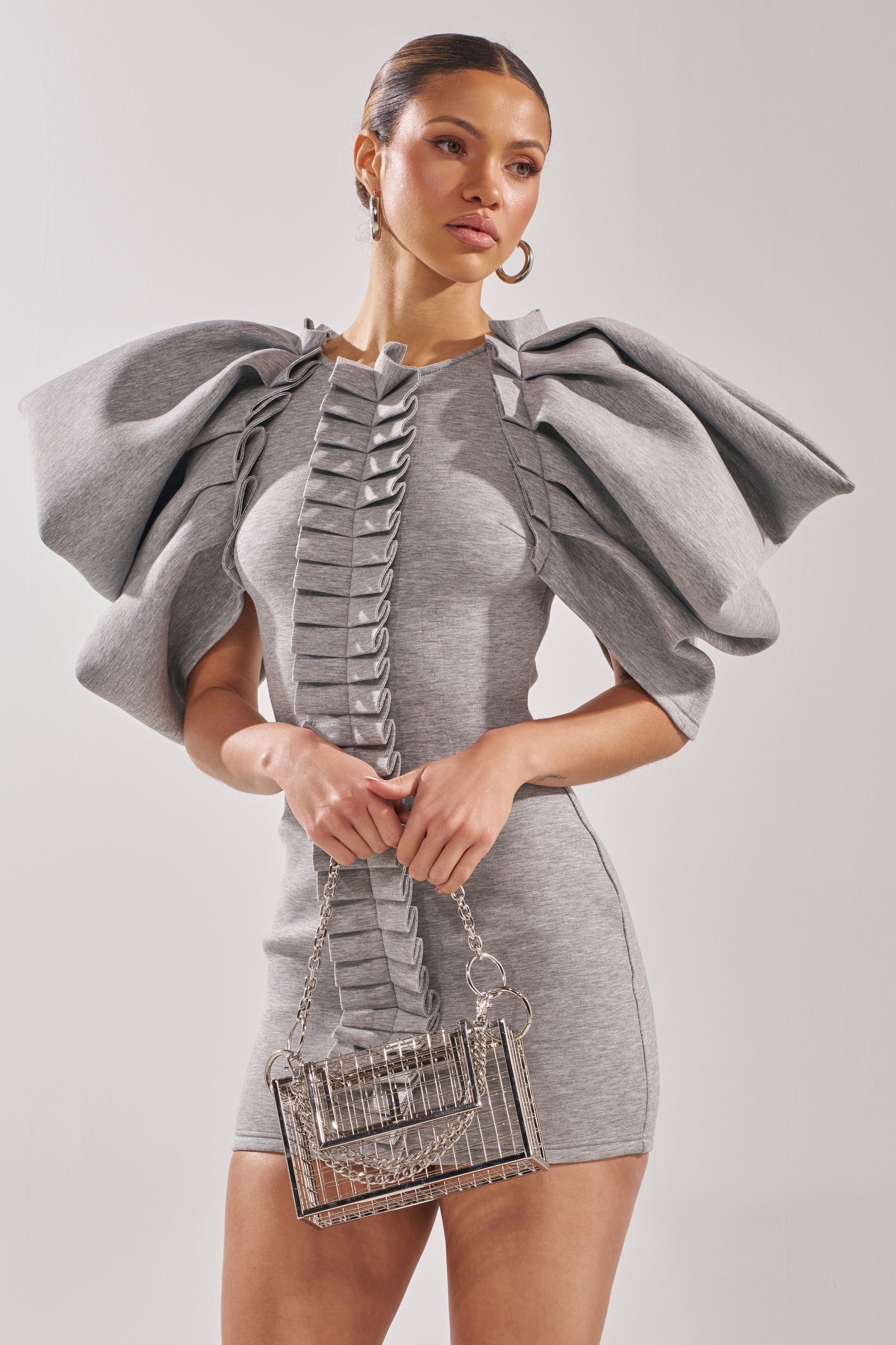 A woman wears the SPREAD MY WINGS MINI DRESS, a fitted heather grey bodycon mini with bold ruffle sleeves, styled with a silver cage handbag and hoop earrings, and her hair is neatly pulled back.