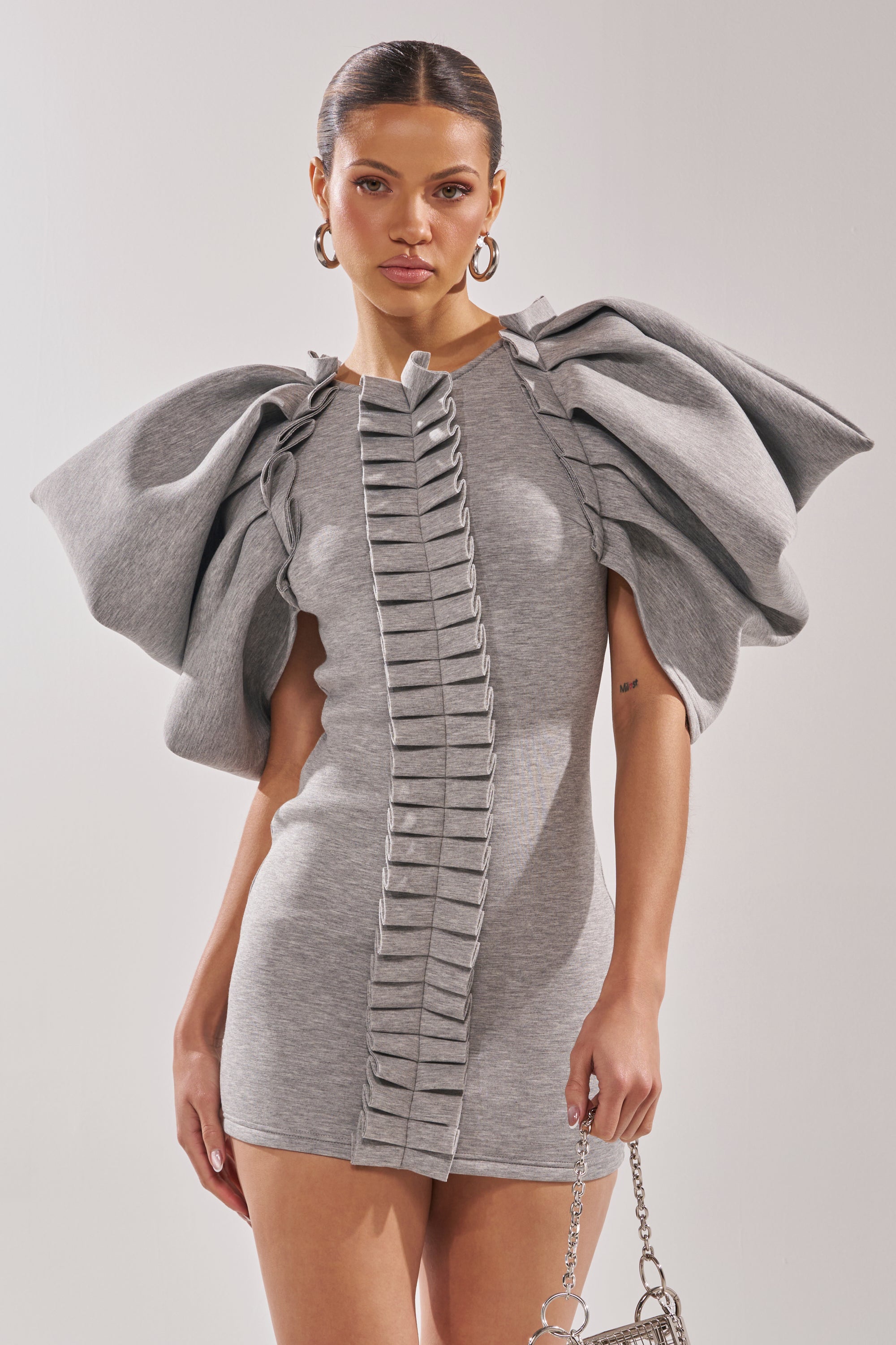 A woman wears the SPREAD MY WINGS MINI DRESS in heather grey, featuring dramatic ruffle sleeves and vertical pleats. She carries a small silver chain handbag and styles her hair in a sleek low ponytail.