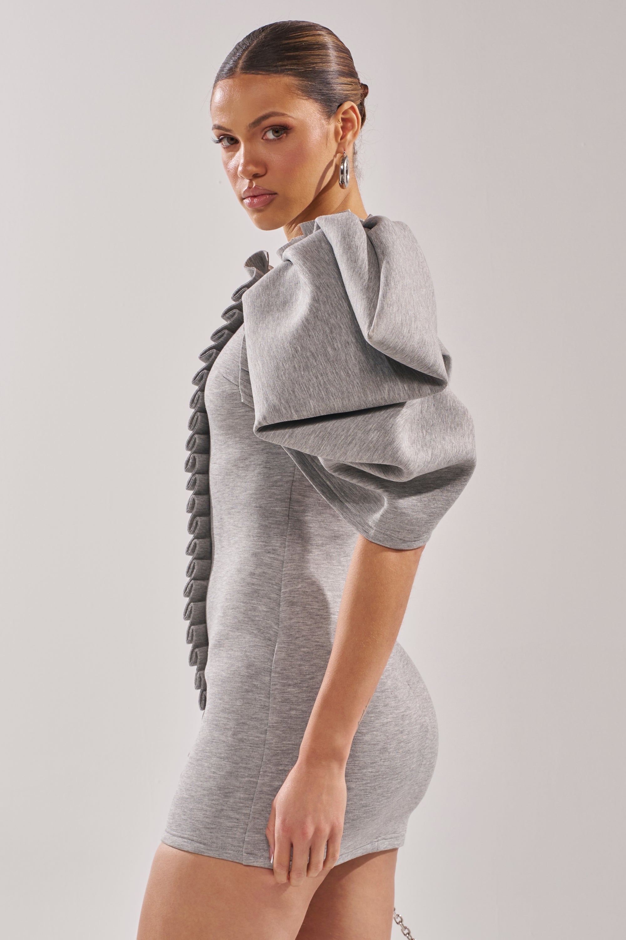 A woman wears the SPREAD MY WINGS MINI DRESS, a fitted heather grey bodycon mini with oversized puff sleeves and button details, standing sideways against a plain light background.