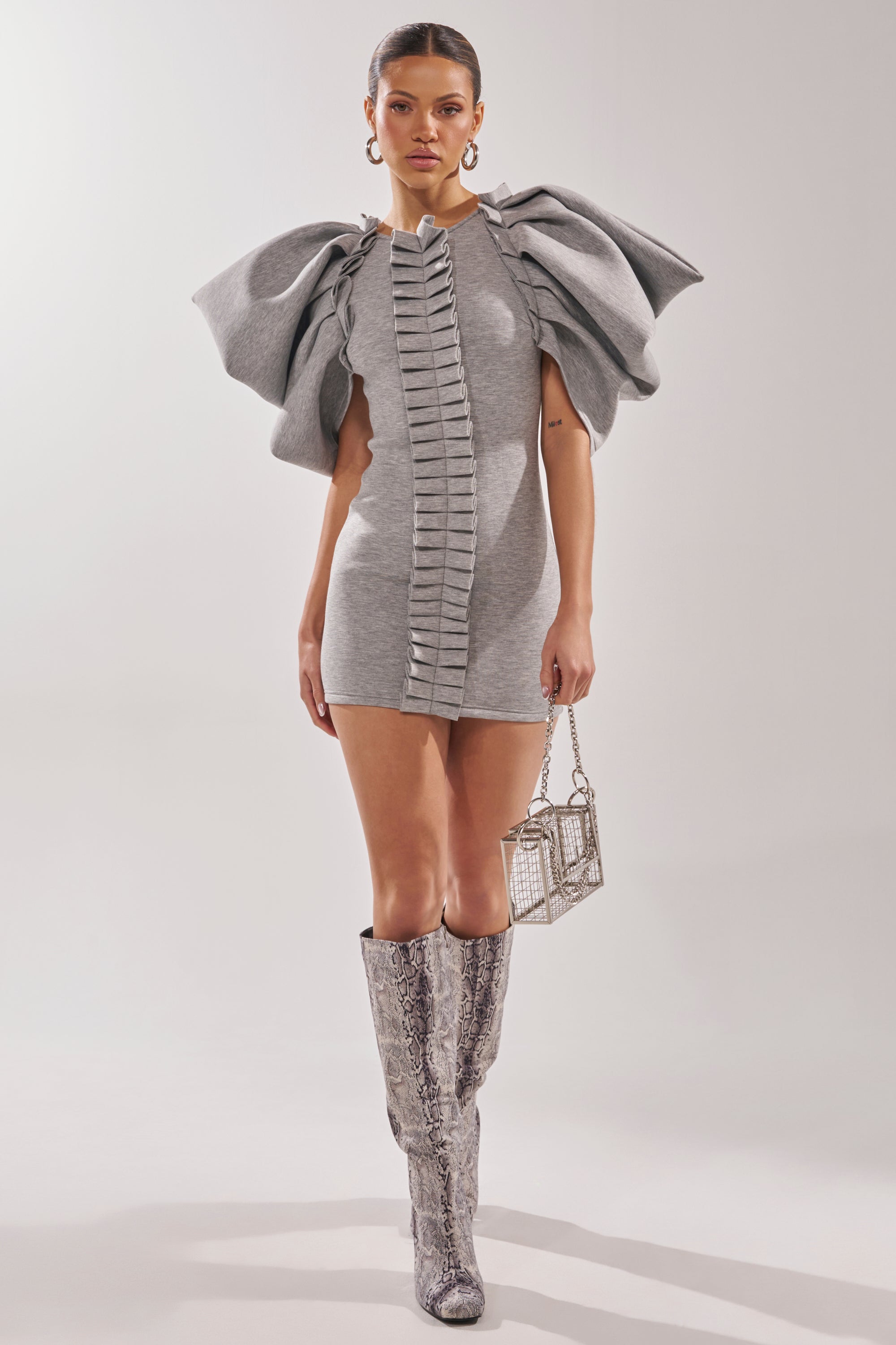 A woman models the SPREAD MY WINGS MINI DRESS in heather grey, featuring ruffle sleeves and a central ruffle. She accessorizes with a small metallic handbag and knee-high snakeskin-patterned boots.